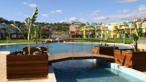 A swimming pool with wooden deck and planters, surrounded by colorful buildings. - Luxury 2-bed apartment with huge pool - Orada resort (Albufeira)