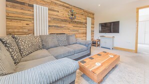 A living room with a sectional sofa, a wooden coffee table, a TV, and a wooden wall panel. - 1 bedroom accommodation in Ulverston (Ulverston)