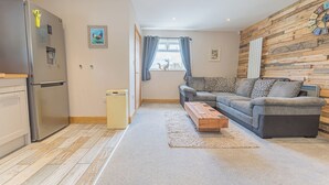 A modern living room with a sofa, a wooden accent wall, a refrigerator, and a window with curtains. - 1 bedroom accommodation in Ulverston (Ulverston)