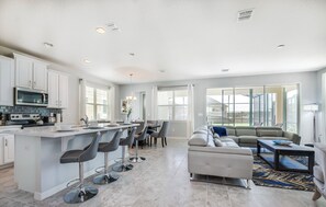 Cocina moderna con armarios blancos, una isla central con taburetes y un comedor con sofá gris y mesa de centro.