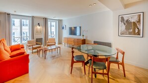 A modern living room with a glass dining table, orange sofa, and wooden chairs. - Madeleine- Charming, quiet, upscale apartment - 2 bedrooms 6 people (Paris)