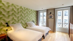 A hotel room with two beds, a green leaf-patterned wall, a window with a view of buildings, and a wooden floor. - Madeleine- Charming, quiet, upscale apartment - 2 bedrooms 6 people (Paris)