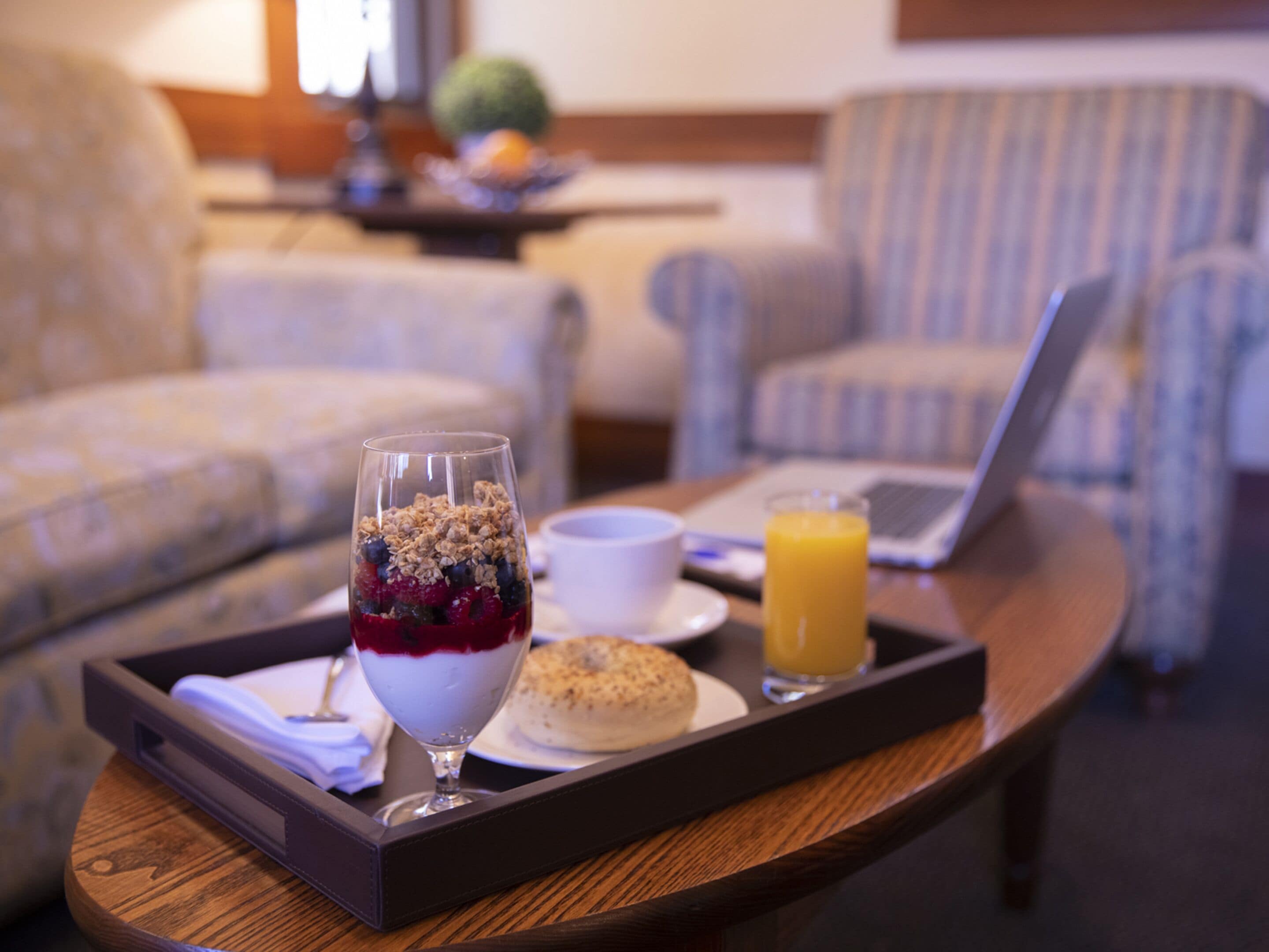 A tray with breakfast items including a glass of yogurt parfait, a muffin, and a glass of orange juice on a wooden table in a hotel room.