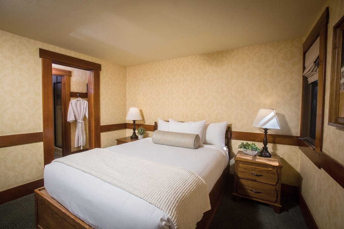 Habitación de hotel con una cama grande, dos mesitas de noche con lámparas, un tocador de madera y una puerta que da al baño.