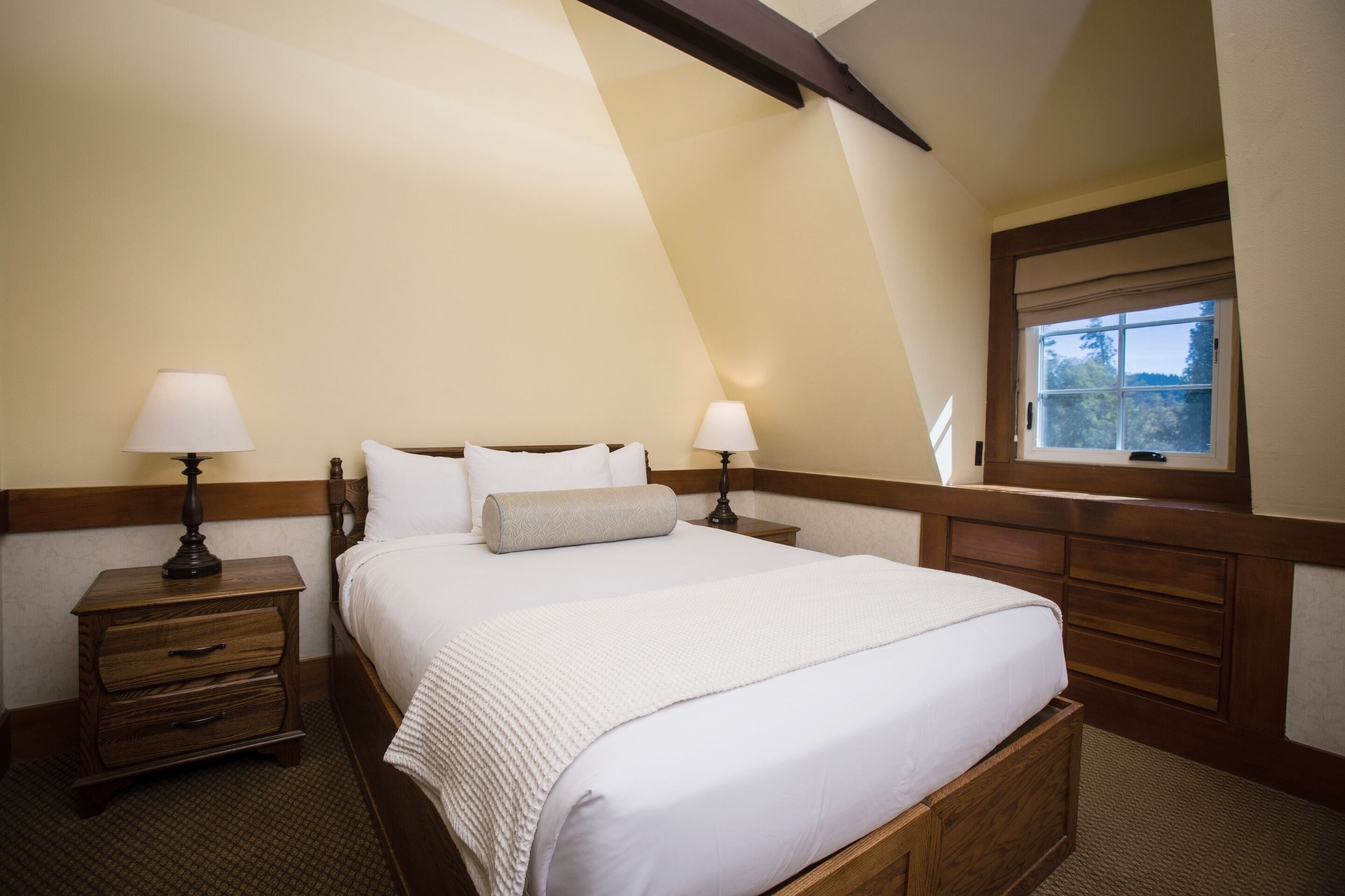A bedroom with a large bed, two bedside tables with lamps, a window with a view, and wooden paneling.