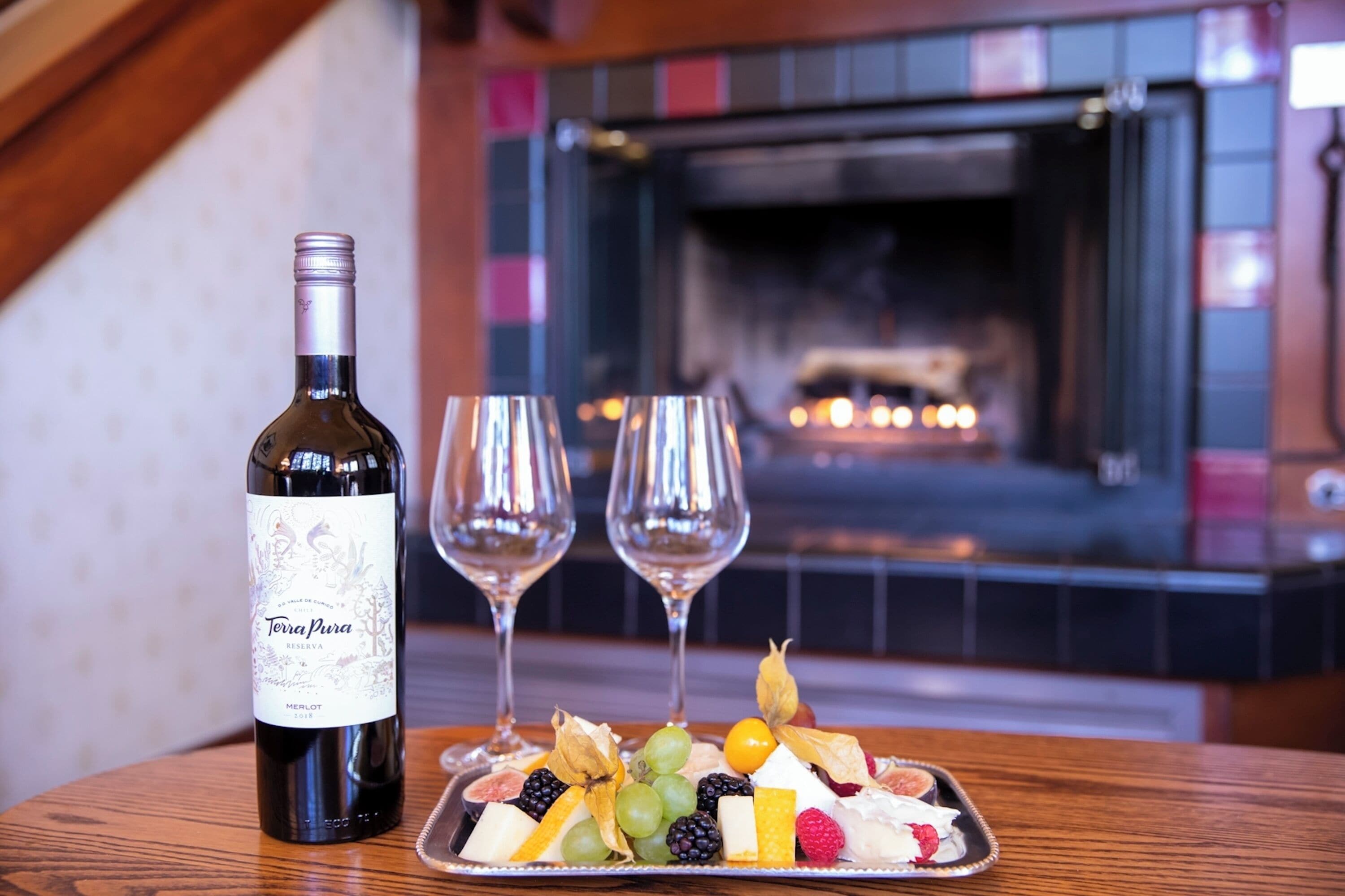 Une bouteille de vin, deux verres et une assiette de fruits sur une table, avec une cheminée en arrière-plan.