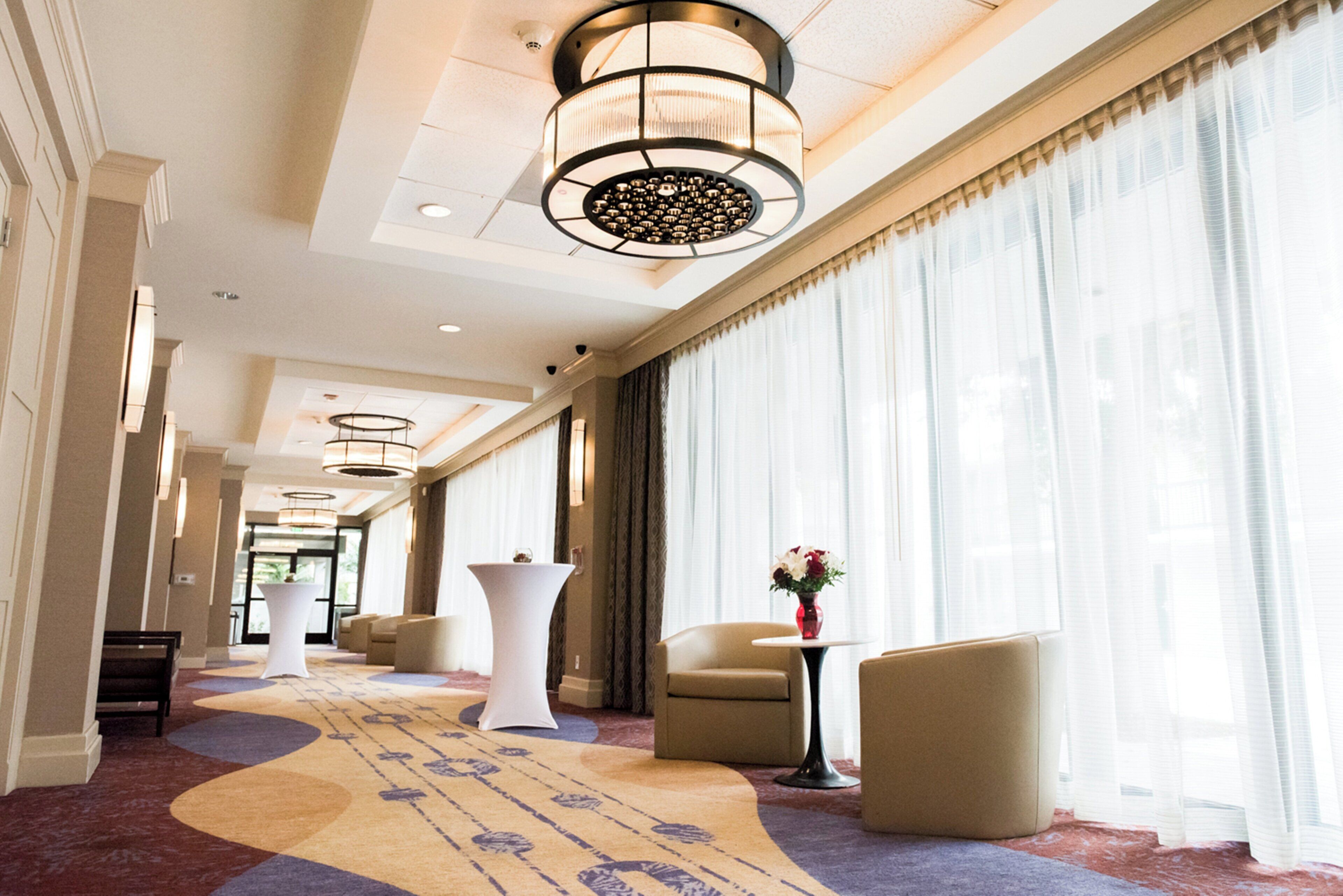 A hotel corridor with a patterned carpet, seating areas, and a large window with sheer curtains.