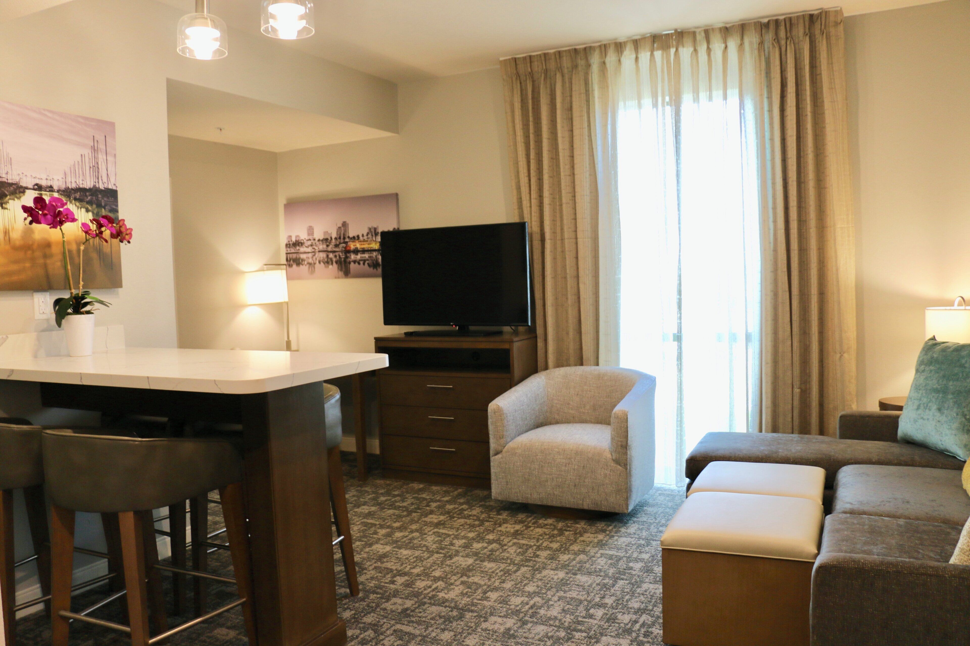 Ein Hotelzimmer mit Küchenzeile, einem Flachbildfernseher, einer Couch, einem Sessel und einem großen Fenster mit Vorhängen.