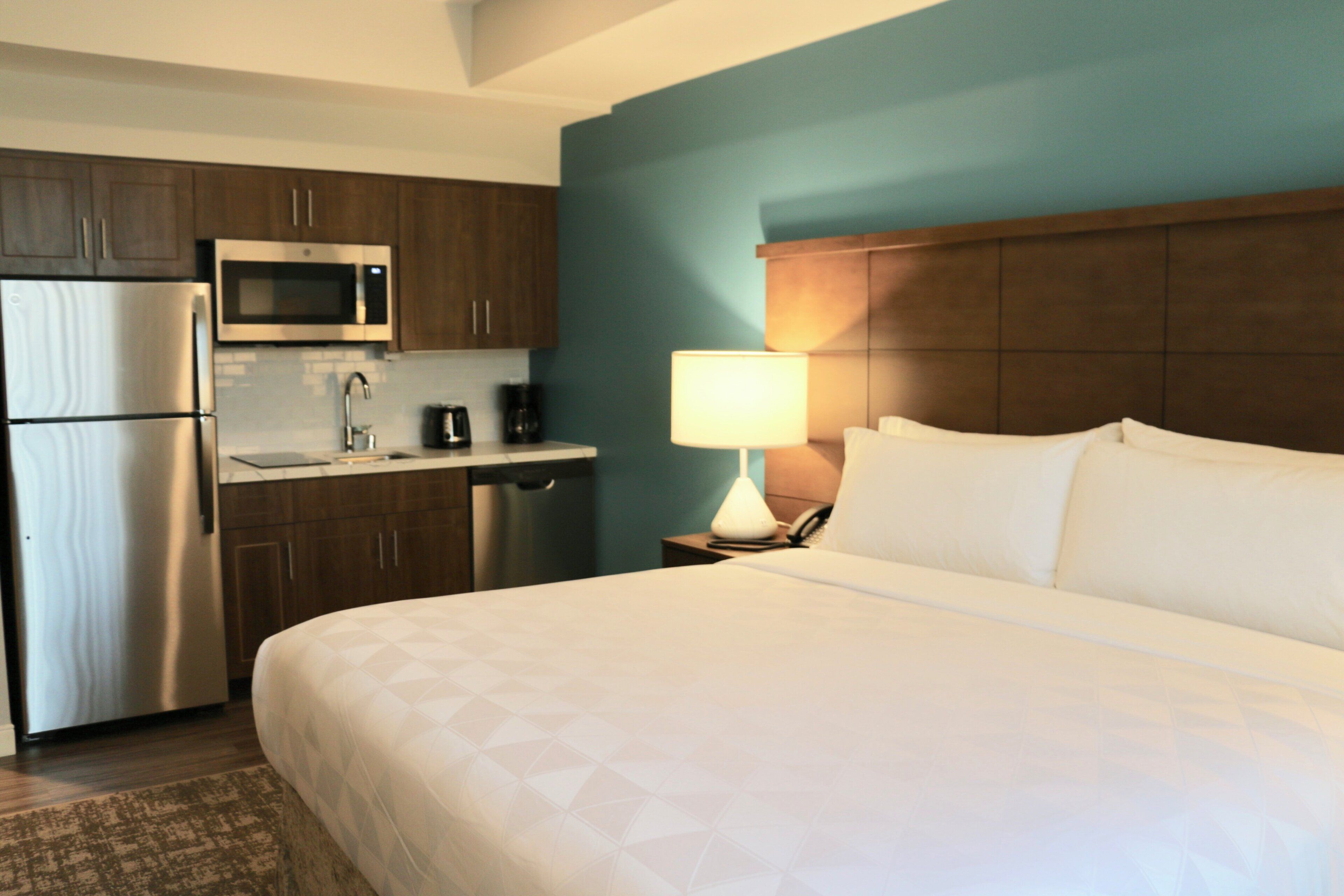 A hotel room with a bed, a nightstand with a lamp, a microwave, a coffee maker, and a refrigerator.