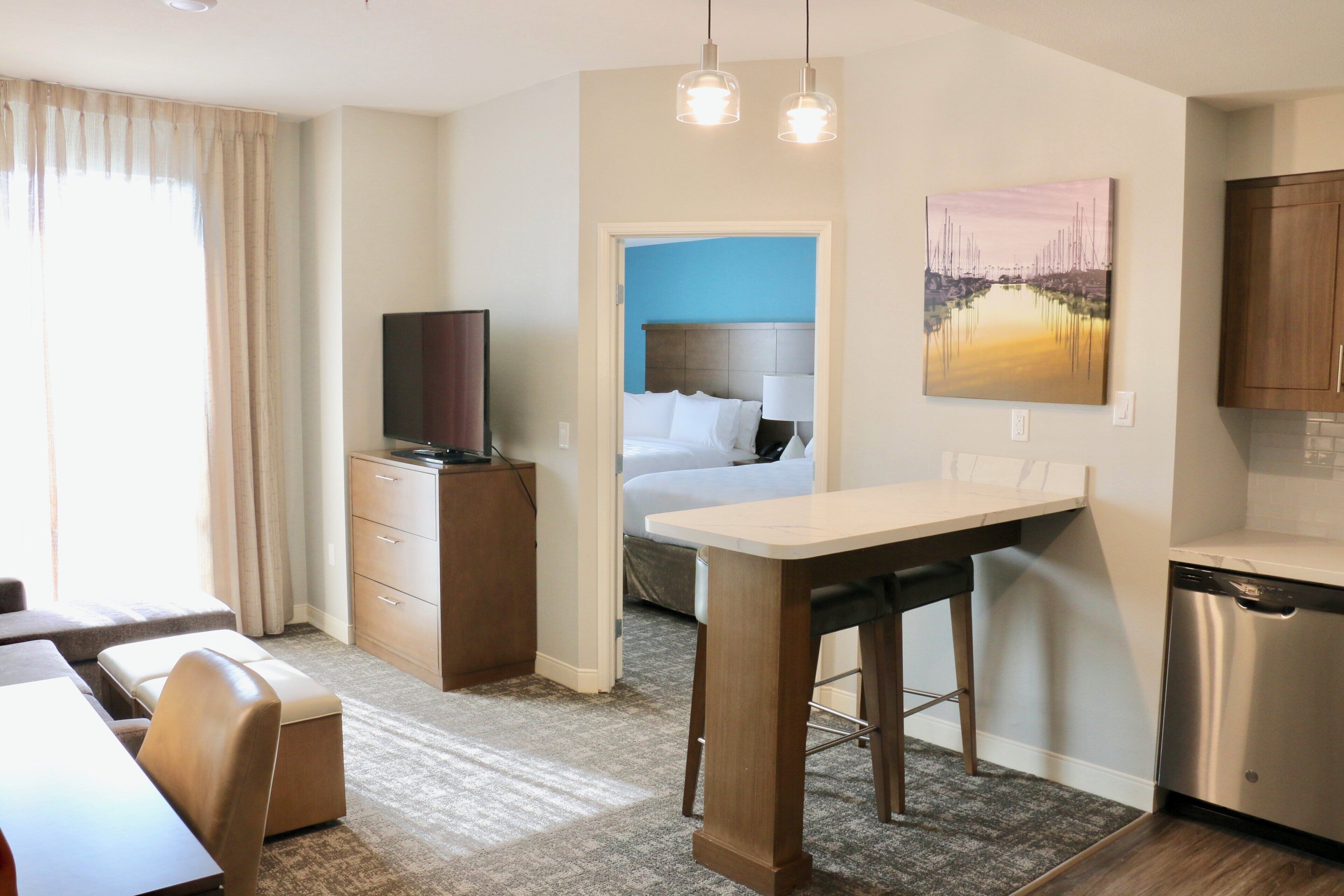 A hotel room with a bed, a desk, a television, a painting, and a kitchen area.