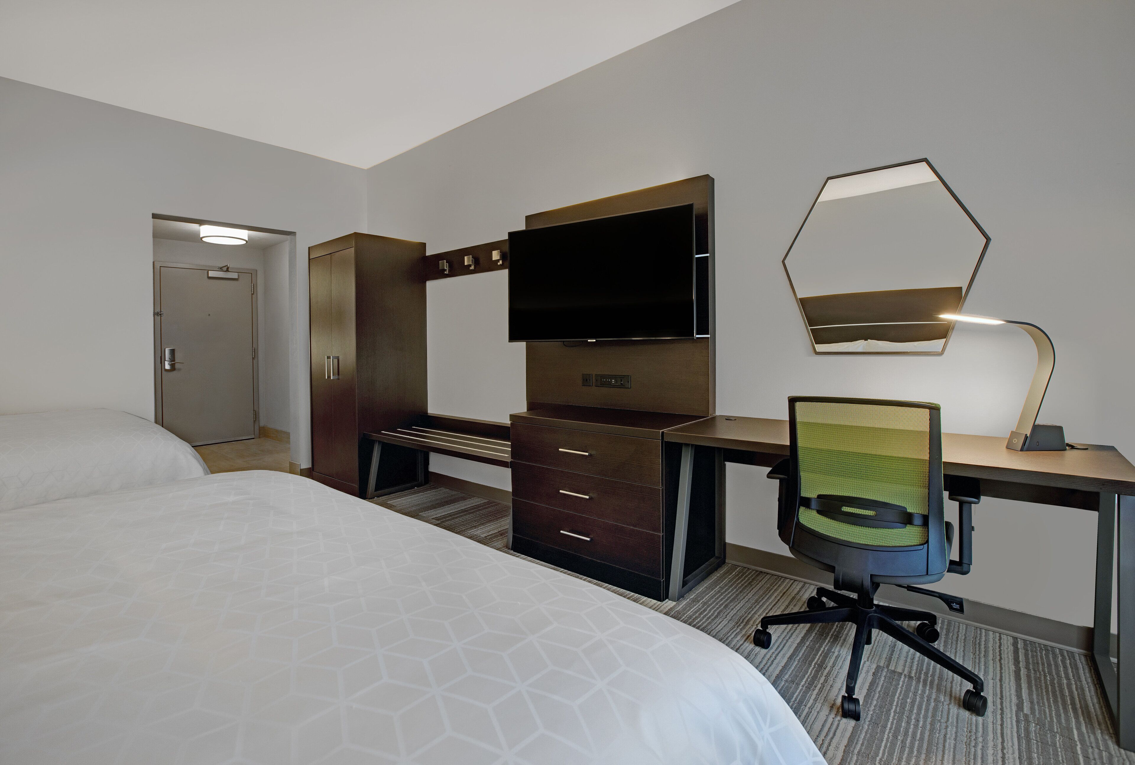 A hotel room with a bed, a desk with a chair, a TV, a mirror, and a wardrobe.