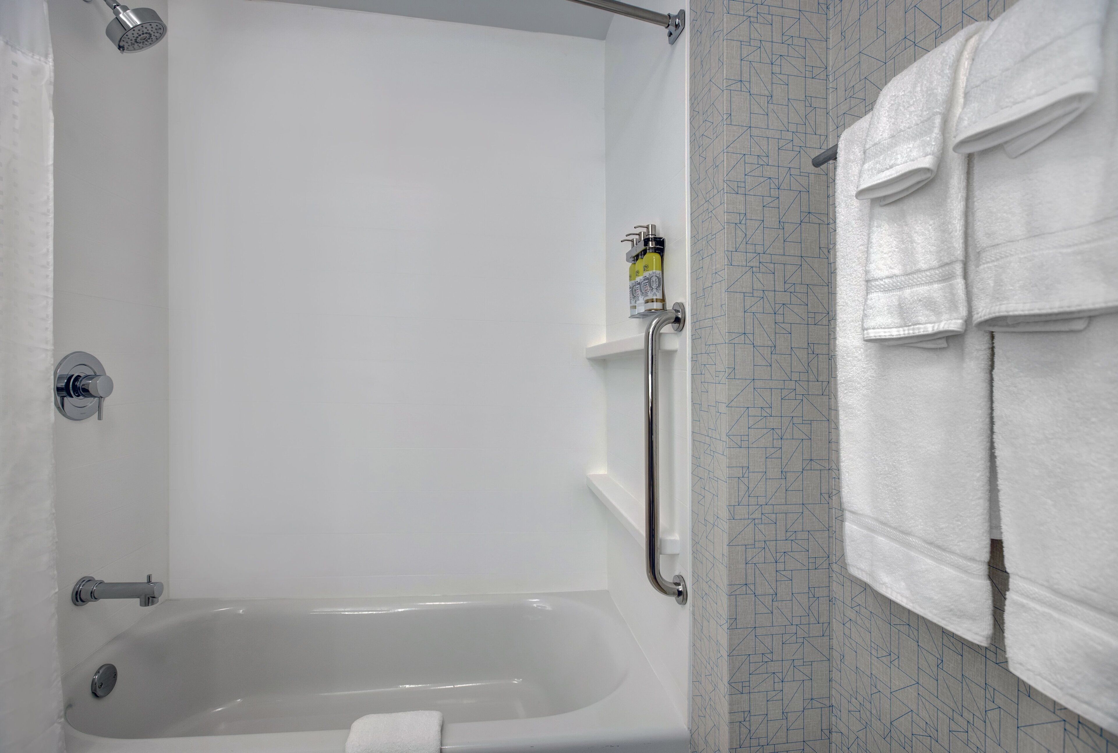 A bathroom with a bathtub, a showerhead, a towel rack, and a soap dispenser.