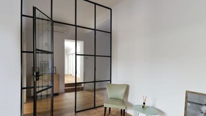 Une pièce dotée d’une porte-fenêtre coulissante donnant sur un autre espace, d’un fauteuil vert clair, d’une petite table ronde avec un plateau en verre et d’un tableau encadré accroché au mur.