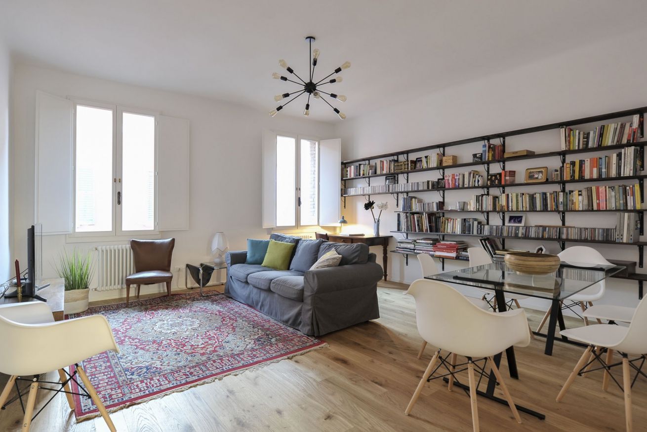 A modern living room with a grey sofa, white chairs, a glass dining table, a bookshelf, and a large window.