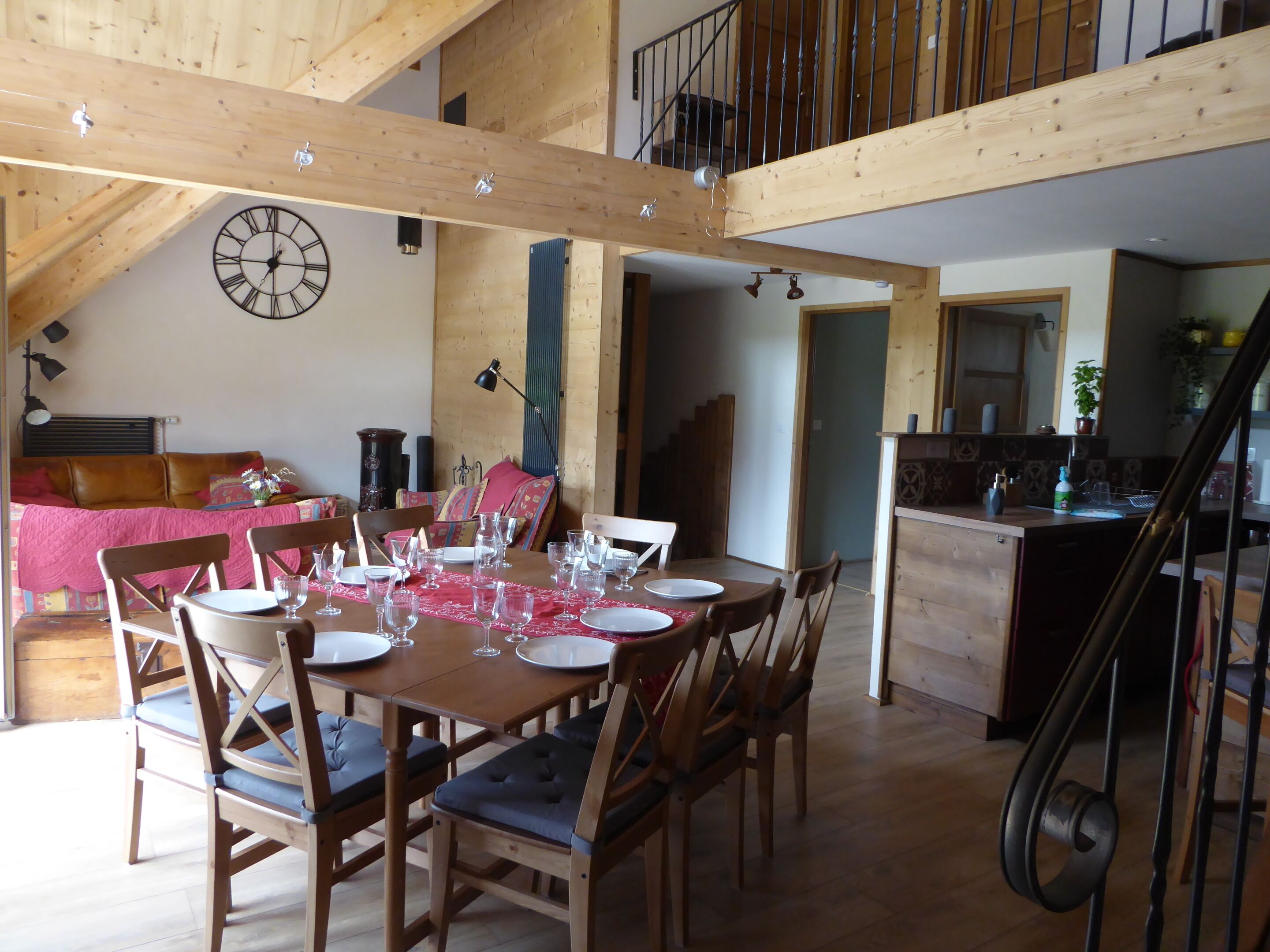 A dining area with a wooden table set for six, surrounded by wooden chairs with cushioned seats. A large wall clock is visible on the wall.