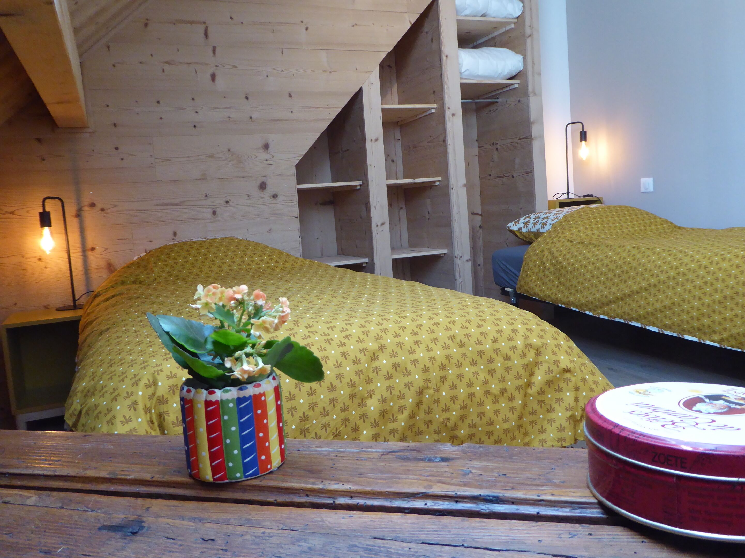 A room with a bed covered in a yellow patterned blanket, a small table with a colorful vase, and a red tin box.