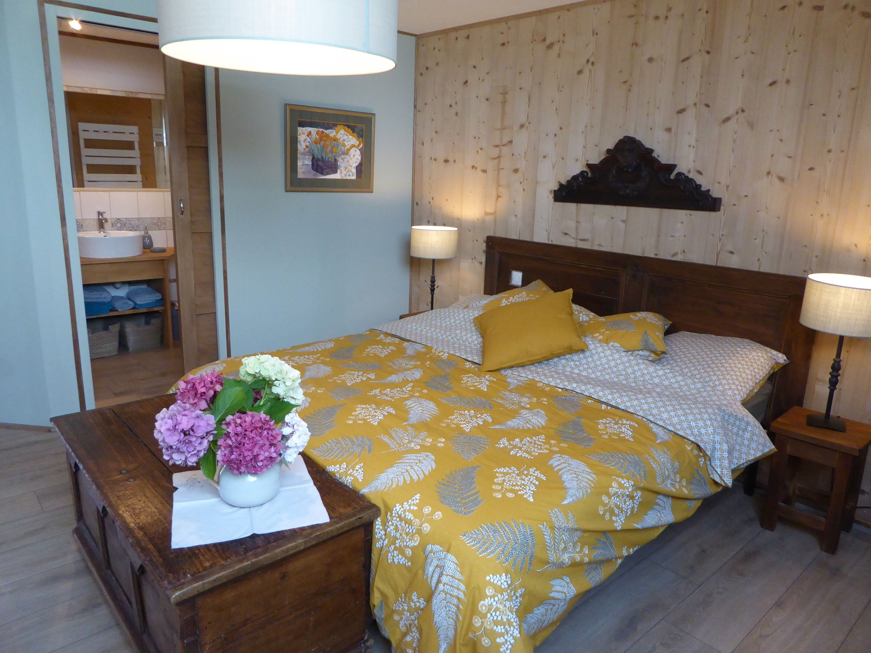 Un dormitorio con cama, mesitas de noche, mesilla auxiliar, cabecera de madera, estante en la pared y baño visible a través de una puerta abierta.