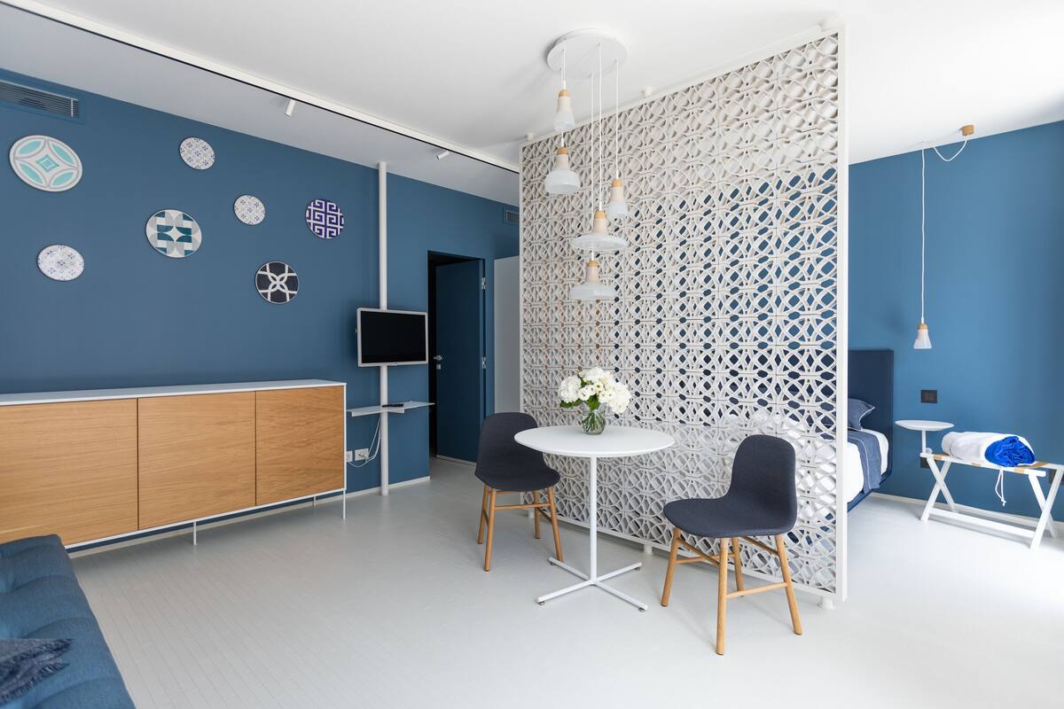A modern room with a blue wall, a white partition, a round table with two chairs, a TV, and a bed.