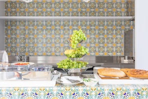 A buffet setup with a tiered fruit display, a bread basket, and various dishes on a table with a patterned tablecloth. - Santa Caterina - Dimora sul mare (Polignano a Mare)