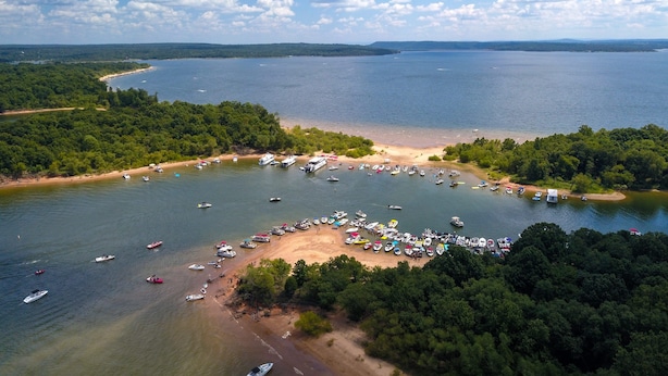 Vue aérienne d’une zone au bord d’un lac, avec des bateaux amarrés le long de la rive et une plage où se trouvent des personnes et des parasols.