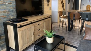 A modern living room with a flat-screen TV, wooden cabinets, a coffee table with a plant, and a dining area with chairs and a kitchen in the background.