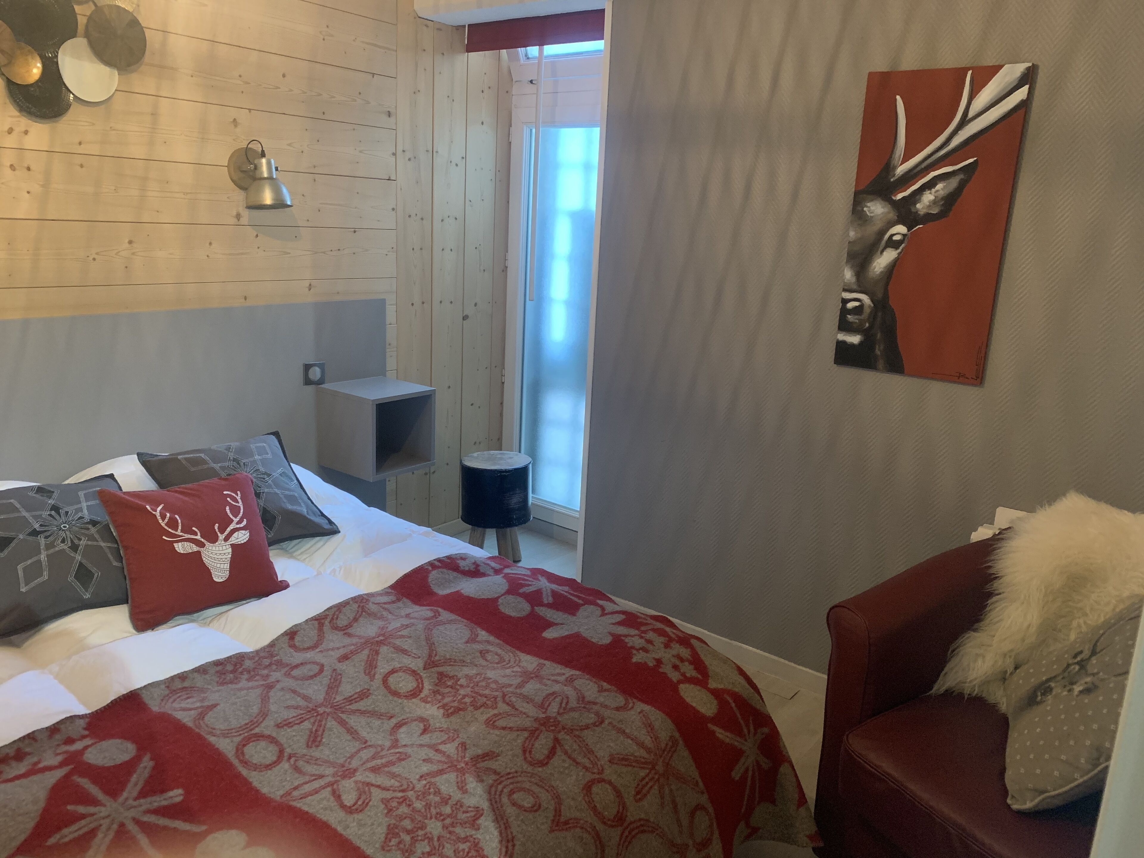 A bedroom with a bed, a red and white patterned blanket, a headboard, a wall-mounted lamp, a small side table, and a painting of a deer.