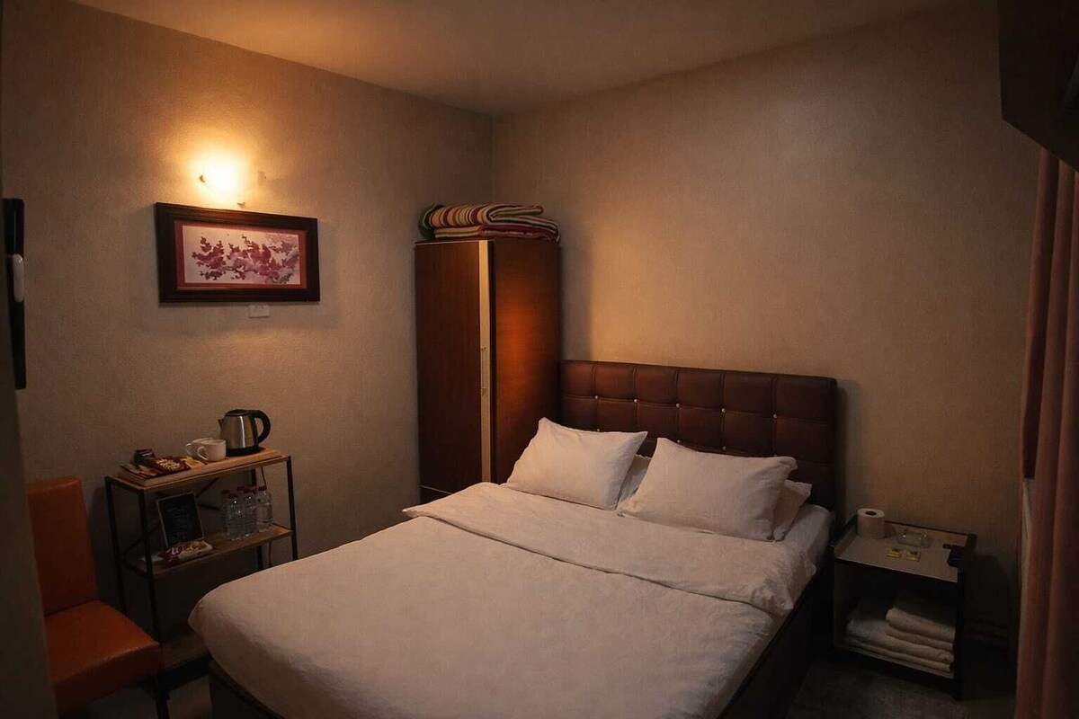 A hotel room with a bed, a nightstand, a shelf with toiletries, a framed picture on the wall, and a tall cabinet.