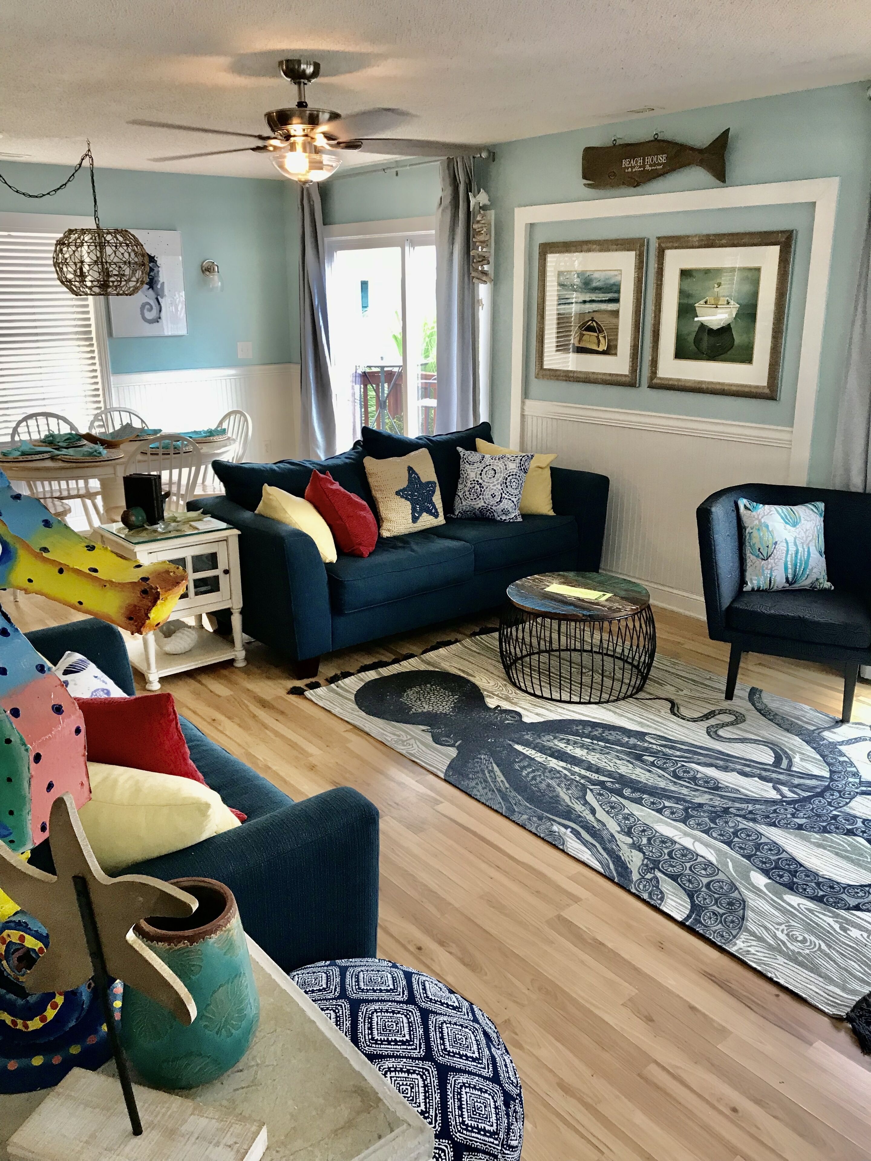Un salon avec un canapé bleu, une table basse et un tapis à motifs. On y trouve également une table à manger avec des chaises et un ventilateur de plafond.