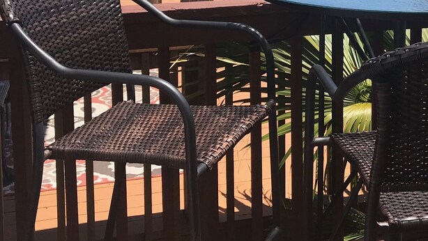 A patio area with wicker chairs and a round table, overlooking a beach with umbrellas and a pool.