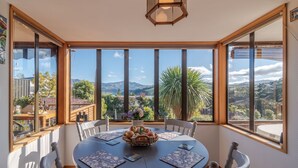 A dining area with a round table, chairs, and a view of the outdoors through large windows. - Pointers - Akaroa Holiday Home (Akaroa)