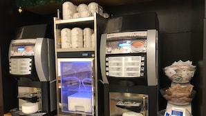 Three coffee machines with a variety of coffee options, a shelf with cups, and a bowl of coffee pods.