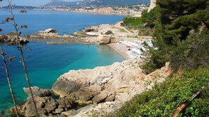 A coastal landscape with rocky terrain, a clear blue sea, and a distant town.