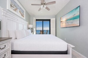 Ein Schlafzimmer mit einem großen Bett, einem Deckenventilator, Blick auf den Ozean durch eine Balkontür und einem Gemälde mit Strandmotiv.