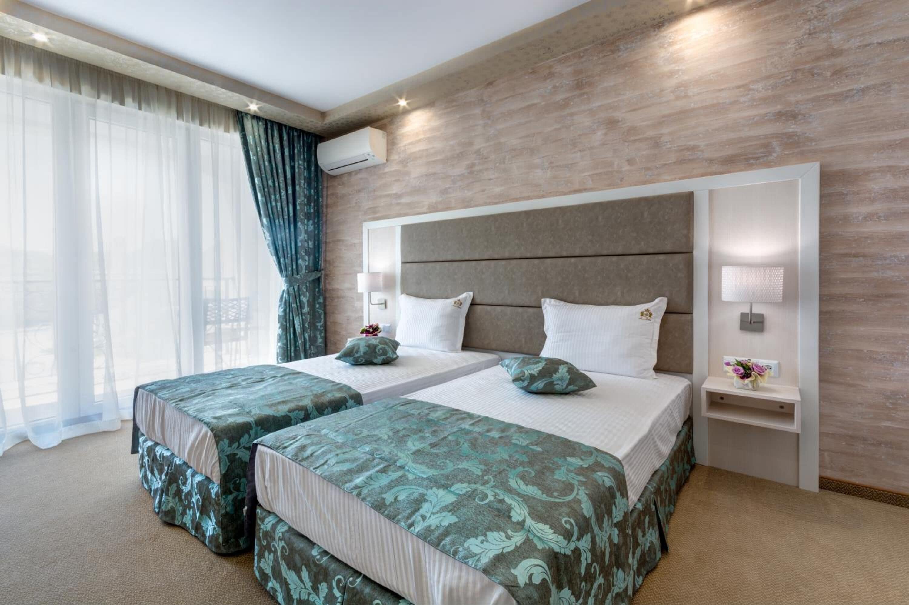 Habitación de hotel con dos camas, un ventanal grande con cortinas translúcidas, un revestimiento de madera y una mesita de noche con lámpara.