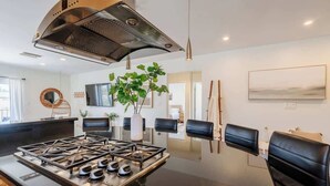 Una cocina moderna con estufa a gas, sillas negras, una planta grande y un cuadro en la pared.