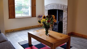Una sala de estar con chimenea, una mesa de madera con un florero, una ventana con contraventanas de madera y un sofá gris.