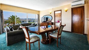 Ein Hotelzimmer mit großem Fenster, einer Couch, einem Esstisch mit Stühlen, einem Spiegel und einer Tür.