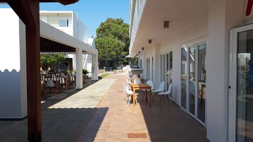 Un patio al aire libre techado con mesas y sillas, un sendero y un edificio con puerta y ventanas de vidrio.