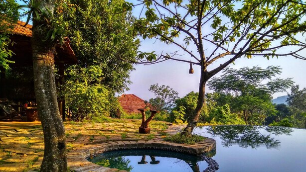 A tranquil outdoor setting with a stone pool, lush greenery, and a traditional thatched-roof building. - Annapara Home Stay (Vythiri)