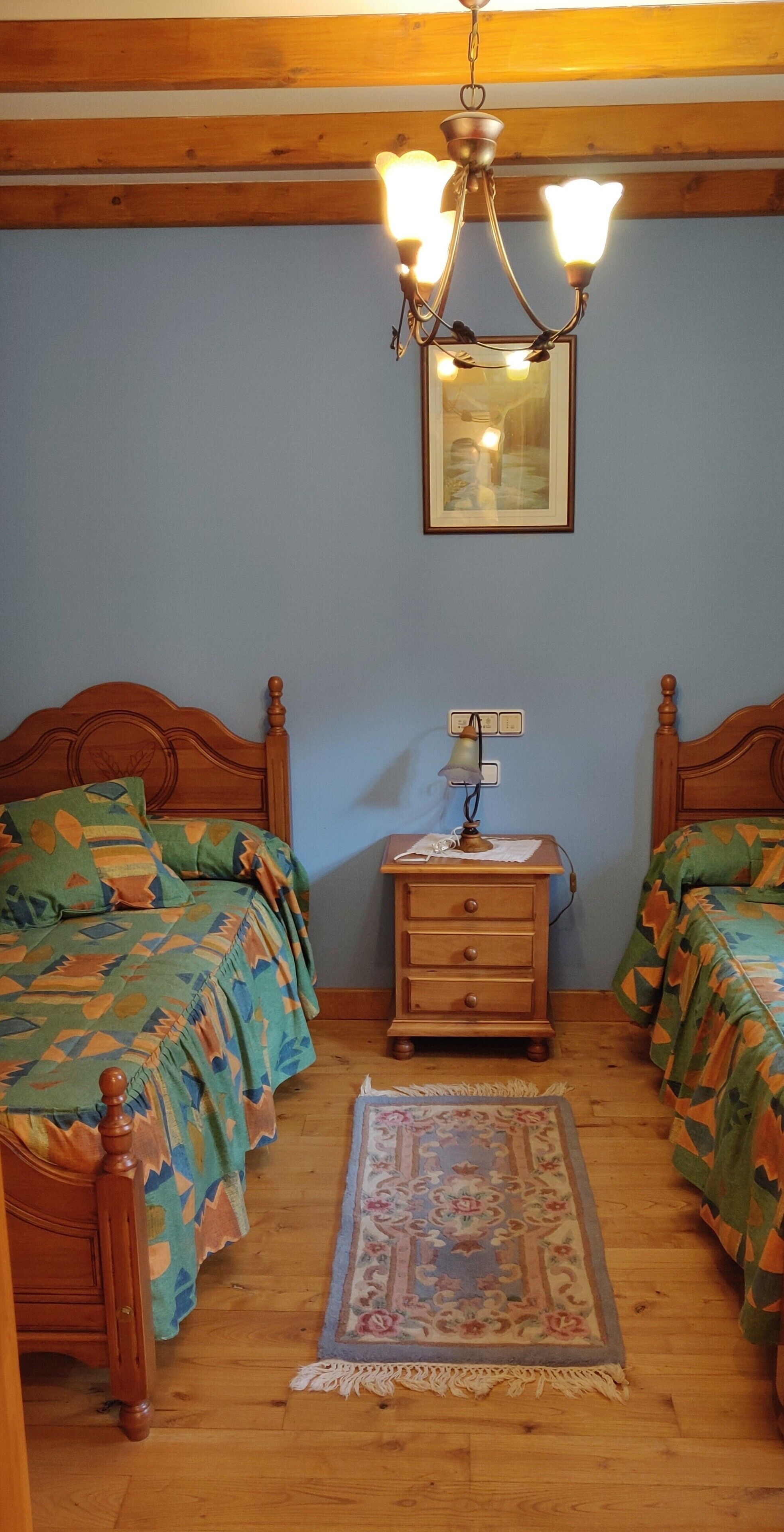 Quarto com duas camas de madeira, uma mesa de cabeceira, um abajur, um candelabro e um quadro na parede.