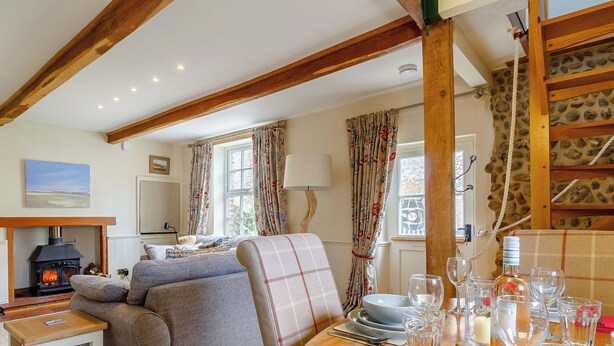 A cozy living room with a fireplace, a sofa, a dining table set for two, and a stone wall. - 2 bedroom accommodation in Blakeney (Blakeney)
