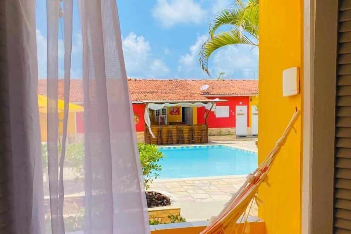 A room with a view of a swimming pool, a hammock, and a red building with a tiled roof.