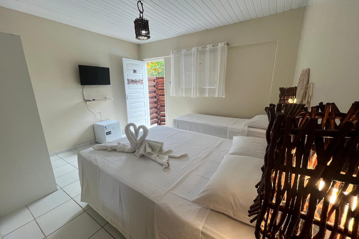 A hotel room with two beds, a TV mounted on the wall, a small white cabinet, and a window with a view of greenery.