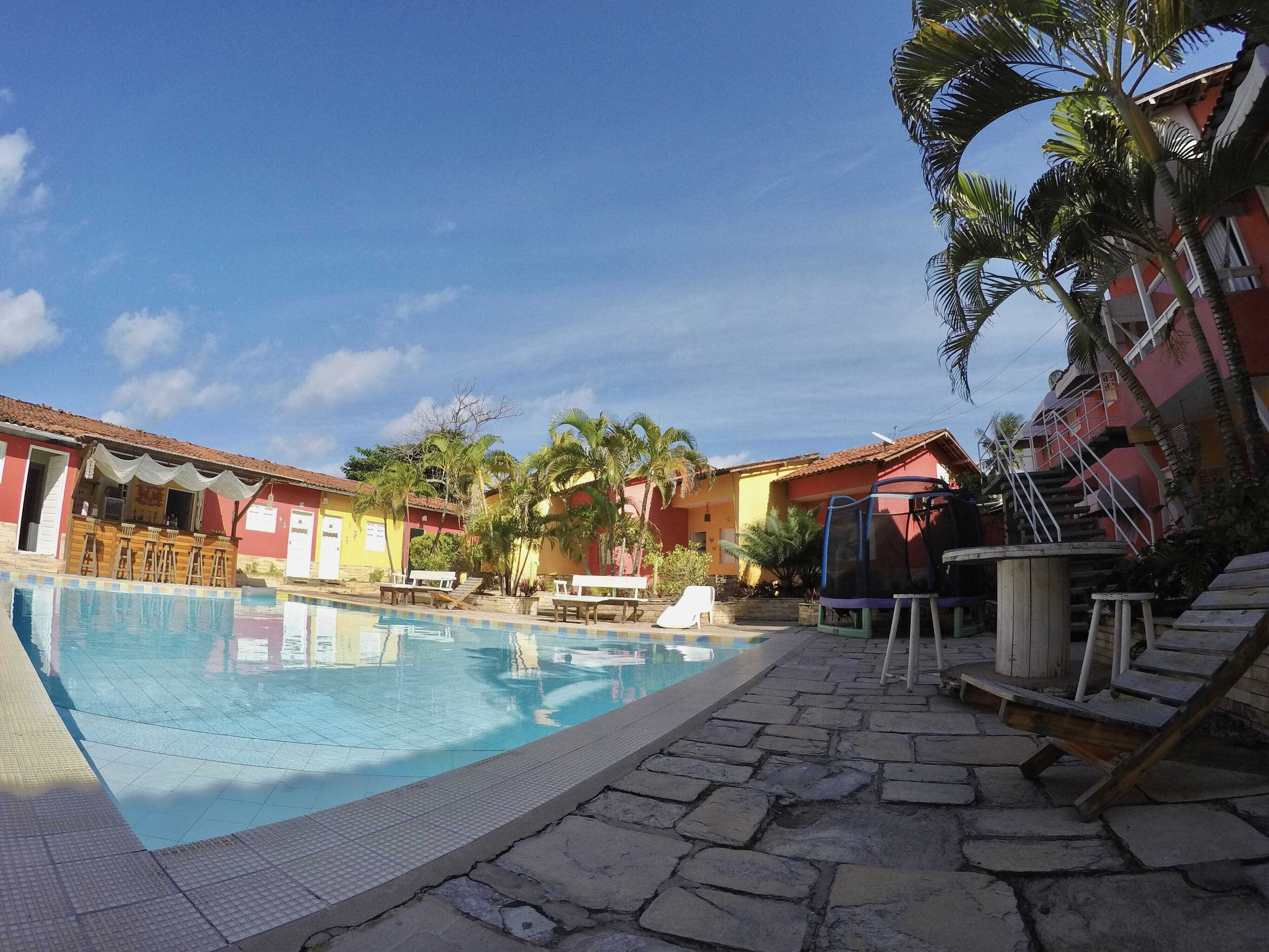A swimming pool with lounge chairs and a bar area, surrounded by palm trees and buildings with red and yellow facades.