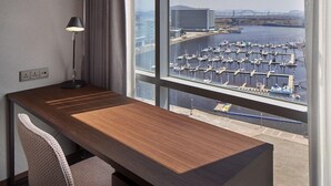 Une chambre d’hôtel avec un bureau en bois, une chaise, une lampe et une grande fenêtre donnant sur un port de plaisance et des immeubles.