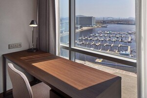 Habitación de hotel con un escritorio de madera, una silla, una lámpara y un amplio ventanal con vistas a un puerto deportivo y edificios.