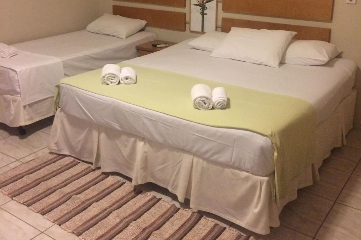Hotelkamer met twee éénpersoonsbedden en één tweepersoonsbed, elk met witte lakens en een groene sprei. Op het tweepersoonsbed liggen twee opgerolde handdoeken. De kamer heeft een houten hoofdbord, een spiegel aan de muur en een gestreept vloerkleed.