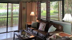A living room with a brown leather sofa, a coffee table, and a lamp. There is a large window with curtains and a view of greenery outside.