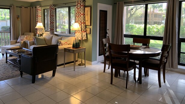 A living room with a sofa, chairs, a coffee table, and a dining table. There are curtains, a lamp, and a ceiling fan.