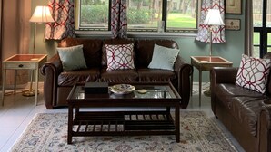 A living room with a brown leather sofa, patterned cushions, a coffee table, and a patterned rug. There are two lamps, a window with curtains, and a framed picture on the wall.